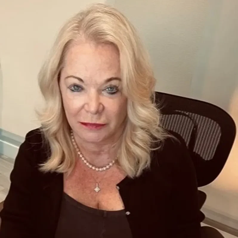 A woman with shoulder-length blonde hair sits in a black office chair, wearing a black top, necklace, and looking up at the camera with a neutral expression.